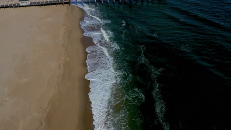 cinematic-aerial-shot-along-the-ocean-beach-edge-tilts-and-lifts-to-reveal-a-dock-pier-and-birds-flying