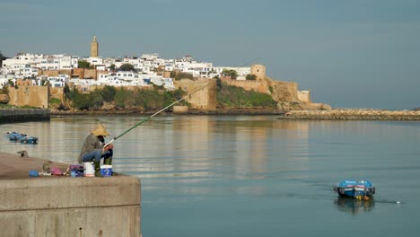 Pescador-En-El-Bou-Regreg-Con-La-Antigua-Ciudad-Y-Fortaleza-De-Rabat,-Marruecos-En-El-Fondo