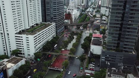 Bangkok-Thailand,-Modern-Metropolis-Midtown-Aerial,-Boulevard-Traffic-Between-Skyscrapers-and-BTS-Skytrain-Train-on-Curvy-Railway