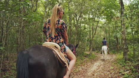 Slow-Motion-Back-shot-of-Young-Blonde-Girl-with-Flower-Dress-Horseback-Riding-inside-Tropical-Forest-Pathway-in-Cancun,-Mexico