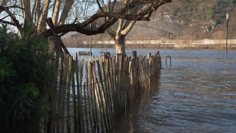 Antigua-Valla-De-Madera,-árboles-Y-Farolas-En-El-Agua-De-La-Inundación-Con-El-Río-Al-Fondo