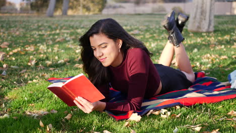 A-young-woman-reading-a-book-in-the-park-during-the-autumn-season-with-fall-leaves-blowing-in-the-wind-as-she-smiles