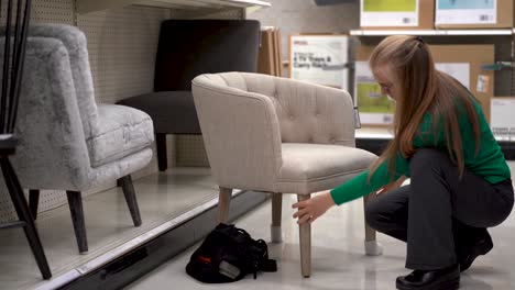 Squatting-down,-a-blonde-woman-scrutinizes-an-upholstered,-modern-looking-chair-in-a-store-display