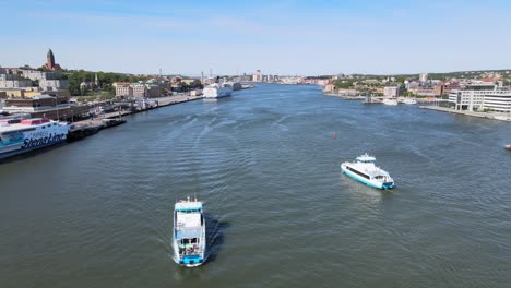Aerial-drone-flight-over-water-with-small-passenger-ferry-crossing-the-Gota-alv-river-and-bigger-ferries-docked-in-harbor-in-Gothenburg,-Sweden-on-a-sunny-day