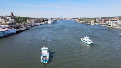 Vuelo-Aéreo-De-Drones-Sobre-El-Agua-Con-Un-Pequeño-Ferry-De-Pasajeros-Cruzando-El-Río-Gota-Alv-Y-Ferries-Más-Grandes-Atracados-En-El-Puerto-De-Gotemburgo,-Suecia-En-Un-Día-Soleado