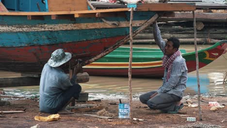 Two-Asian-Men-Fixing-a-Traditional-Wooden-Boat-on-the-Shore