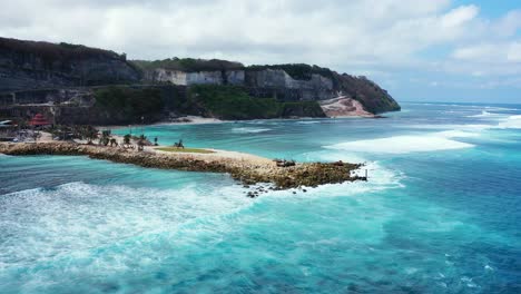 Beautiful-sea-scenery-with-white-waves-splashing-on-rocky-pier-seaside,-slopes-of-green-hills-in-Bali,-Indonesia
