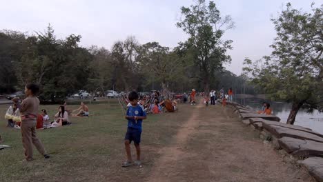 Mittlere-Außenaufnahme,-Die-Sich-Tagsüber-Durch-Menschen-Bewegt,-Die-Sich-Am-Grasbewachsenen-Ufer-Des-Angkor-Grabens-Mit-Bäumen-Entspannen