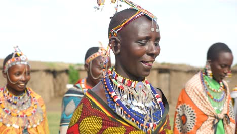 Mujeres-Maasai-Tribales-Bailando-Y-Cantando-En-Primer-Plano,-Con-Todas-Sus-Joyas-Tradicionales-En-El-Fondo-Son-Las-Casas-De-Barro-Que-Usan-Para-Vivir-En-El-Parque-Nacional-Masai-Mara-En-Kenia-áfrica