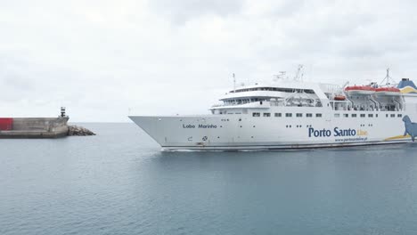 Large-white-passenger-cruise-boat-enters-Porto-Santo-port-waters-by-cement-pier-in-harbor,-Portugal,-handheld-profile-close-up-pan-left