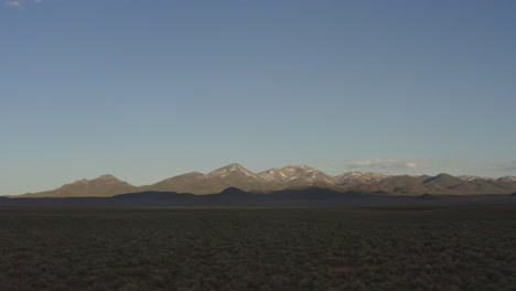Forward-tracking-over-sagebrush-field-at-sunset,-approaching-far-off-snowy-mountains