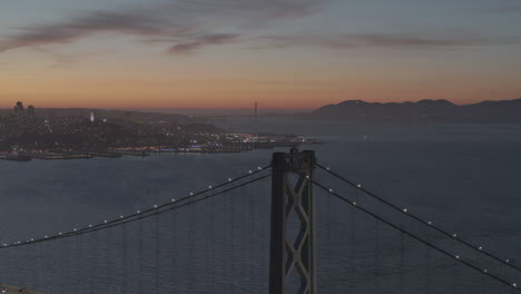 Un-Puente-Colgante-Está-Iluminado-Por-Luces-Mientras-El-Sol-Se-Pone-Sobre-Una-Ciudad-Cercana