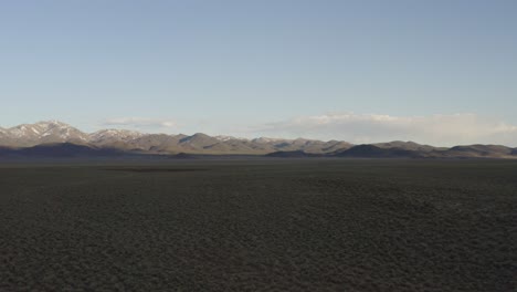 Wide-sagebrush-valley,-tracking-drone-approaching-far-away-snowy-mountain-peaks