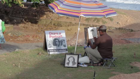 Sketch-Artist-in-Galle-Fort-Perform-a-live-pencil-Portraiture-drawing-of-a-small-child-on-a-piece-of-white-paper-while-using-his-mobile,-sitting-on-a-small-foldable-chair-under-a-big-colorful-umbrella