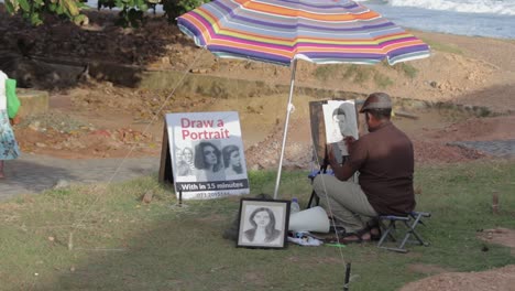 Dibujante-En-El-Fuerte-De-Galle-Realiza-Un-Retrato-A-Lápiz-En-Vivo-De-Un-Niño-Pequeño-En-Un-Trozo-De-Papel-Blanco-Mientras-Usa-Su-Móvil,-Sentado-En-Una-Pequeña-Silla-Plegable-Bajo-Un-Gran-Paraguas-Colorido