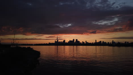Dark-clouds-rolling-into-a-beautiful-sunset,-with-Toronto-skyline-silhouetted-against-the-horizon,-reflecting-on-the-lake