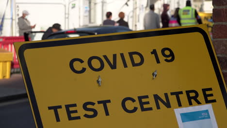People-exit-a-car-and-join-others-in-protective-face-masks-queuing-up-outside-an-appointment-only-Covid-19-testing-centre-behind-a-yellow-Covid-19-Testing-Centre-street-sign
