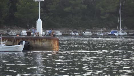 Zwei-Männer-Fischen-An-Einem-Pier-In-Vela-Luka-In-Kroatien,-Als-Ein-Freund-In-Einem-Boot-Vorbeifährt