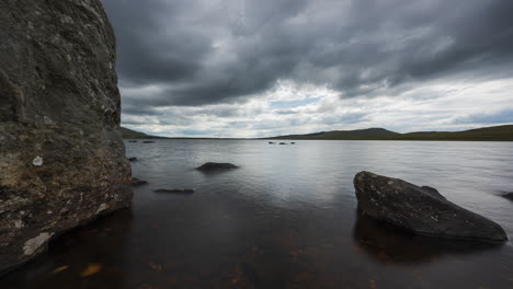 Panorama-motion-time-lapse-of-lake-with-grass-and-large-rocks-in-the-foreground-on-a-dark-cloudy-summer-day-in-rural-landscape-of-Ireland