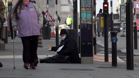 A-man-wearing-a-blue-protective-face-mask-sits-on-the-ground-begging-for-money-with-an-empty-coffee-cup-during-the-Coronavirus-pandemic-in-Winter-as-people-walk-past-ignoring-him