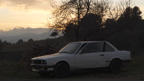 Mann-Fährt-Mit-Weißem-Oldtimer-In-Die-Sonnenuntergangslandschaft,-Parkt-Und-Steigt-Aus