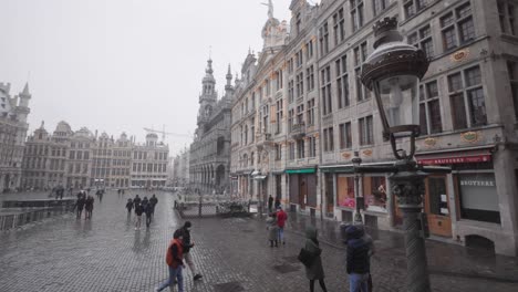 People-walking-through-the-Grand-Place-in-downtown-Brussels,-Belgium-while-snowing,-wide-panning-right