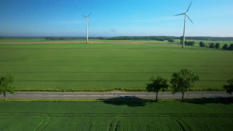 Aerial-trucking-shot-of-electric-car-driving-on-rural-road-and-spinning-wind-turbines-in-background---Beautiful-sunny-day-with-blue-sky-in-countryside
