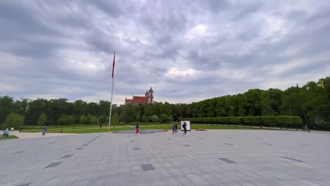 Lithuanian-flag-hanging-on-flagpole-and-people-walking-at-Lukiskes-square-in-Vilnius,-Lithuania