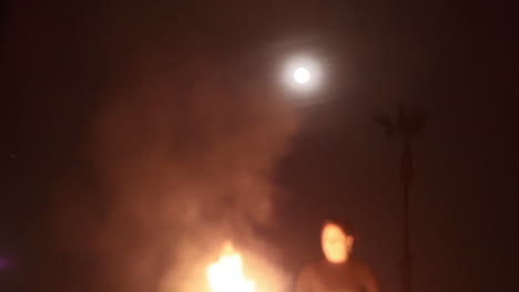 Woman-Builds-Bonfire-at-the-Beach-at-Night,-Palm-Tree-and-Moon,-Out-of-Focus
