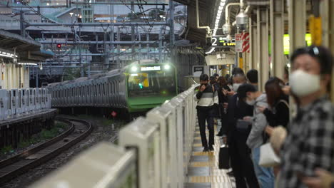 Los-Viajeros-Locales-Con-Mascarilla-Se-Paran-En-La-Plataforma-Con-La-Línea-Yamanote-Acercándose-En-La-Estación-De-Tren-En-Tokio,-Japón