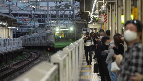 Local-Commuters-Wearing-Face-Mask-Stand-By-At-Platform-With-Approaching-Yamanote-Line-In-Train-Station-At-Tokyo,-Japan