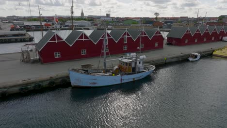 Aerial-view,-old-wooden-ship-is-moored-in-the-port-at-the-quay,-on-the-pier-stand-the-red-wooden-fishermans-warehouses