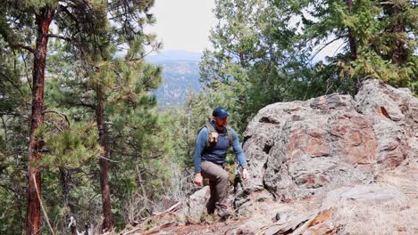 Solo-male-hiker-emerging-from-behind-rocks-and-hiking-through-a-pine-forest-uphill-and-to-the-right-of-the-camera