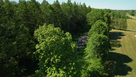Flying-over-a-Countryside-Road-Next-to-a-Green-Field-and-Forest-Trees-Park,-Woodland-and-Landscape-of-Rural-Area-in-Poland-at-Beautiful-Summer-Day