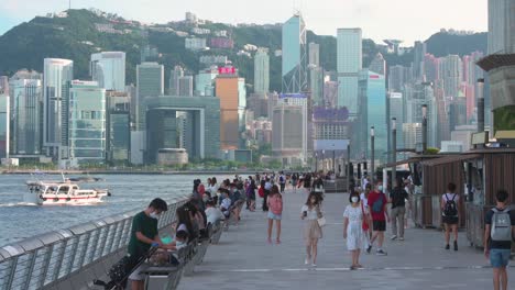 People-enjoy-their-evenings-at-the-Avenue-of-Stars-promenade,-Victoria-Harbour-waterfront,-as-the-city's-skyscrapers-and-skyline-are-seen-in-the-background-in-Hong-Kong