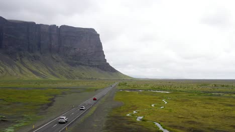 Three-cars-driving-on-Ring-Road-in-Iceland-by-a-mountain-with-drone-video-following-behind