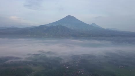 Mystische-Luftaufnahme-über-Einer-Mit-Nebel-Bedeckten-Tropischen-Landschaft-An-Einem-Sonnigen-Morgen-Und-Einem-Bewölkten-Tag-–-Im-Hintergrund-Ragt-Ein-Gewaltiger-Berg-Empor