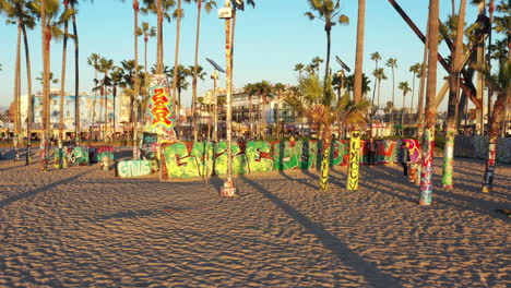 Drone-shot-of-venice-beach-boardwalk-during-sunset-showing-palm-trees,-graffiti-walls,-skateboarding-and-people-taking-photos