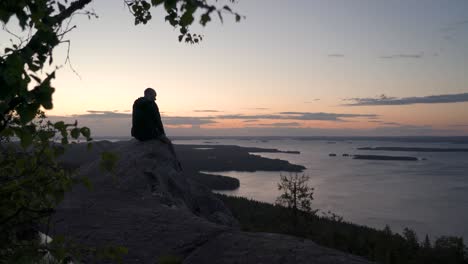 Male-hiker-sitting-on-a-nordic-mountain-cliff-looking-towards-horizon-and-enjoying-a-summer-sunset-or-a-sunrise-over-a-big-lake-view