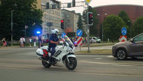 Latvian-road-police-officer-on-the-motorbike-with-blue-and-red-flashing-lights,-monitoring-city-traffic,-summer-evening,-golden-hour,-medium-handheld-tracking-shot