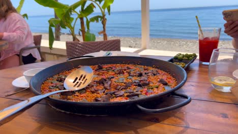 Paella-Tradicional-Española-Con-Pollo-Y-Verduras-En-Un-Restaurante-Junto-A-La-Playa-Con-Vistas-Al-Mar,-Comiendo-Aceitunas-Verdes,-Sabrosos-Mariscos-En-Marbella-España,-Toma-De-4k