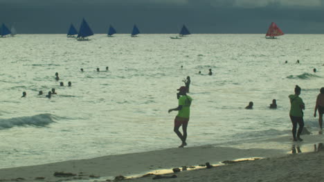 A-lifeguard-on-neon-walks-around-as-tourists,-sail-boats,-and-Boracay-sunset-pass-by