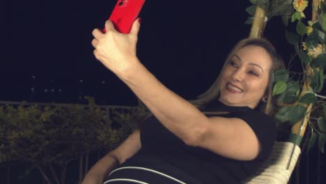 Lächelnde-Hispanische-Frau,-Die-Ein-Selfie-Macht,-Während-Sie-Sich-Nachts-Auf-Einem-Schaukelstuhl-Zurücklehnt