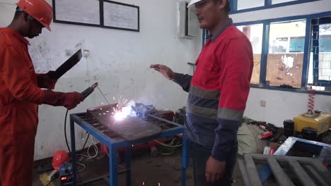 Soldador-Industrial-Con-Antorcha-Y-Casco-Protector-En-Una-Gran-Sala-Soldando-Perfiles-Metálicos-Y-En-Segundo-Plano-Un-Ingeniero-Revisando-El-Plano