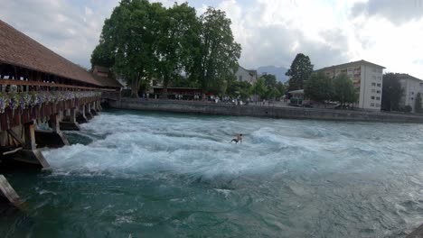 Young-man-surfing-on-Aare-river-by-historical-wooden-bridge-in-Thun,-Switzerland