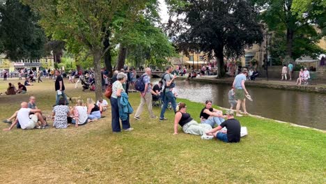 Multitud-De-Personas-Relajándose-En-El-Césped-De-Verano-Junto-Al-Río-En-Bourton-On-The-Water,-Cotswolds---Inglaterra,-Reino-Unido.