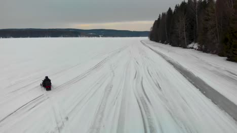 Toma-Aérea-De-Seguimiento-De-Un-Hombre-Conduciendo-Su-Moto-De-Nieve-En-Indalsalven-En-Timra,-Sundsvall,-Suecia