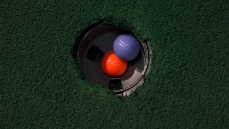 Una-Vista-De-Pájaro-De-Cerca-De-Una-Pelota-Naranja-En-Un-Hoyo-Y-Luego-Una-Pelota-De-Golf-Morada-Y-Amarilla-Cae-En-El-Hoyo-De-Golf-Una-Encima-De-La-Otra