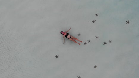 Toma-Aérea-De-4.000-Metros-De-Una-Joven-Flotando-Boca-Arriba-Cerca-De-La-Playa-Rodeada-De-Estrellas-De-Mar