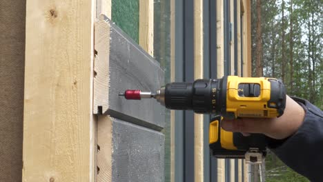 Mano-Masculina-Sosteniendo-Un-Destornillador-Eléctrico-Y-Atornillando-Tablones-De-Madera-Entrelazados-En-La-Fachada-De-La-Casa,-Lengüeta-Y-Ranura